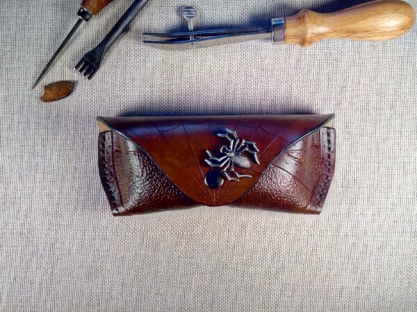 Skórzane etui na okulary na którym wytłoczona jest pajęczyna i siedzi na niej metalowy pająk.